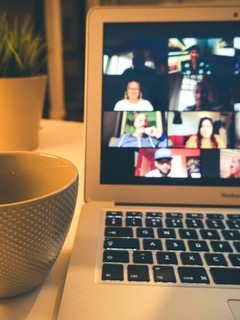 Na zdjęciu widoczny jest laptop stojący na biurku, na którego ekranie wyświetla się wideokonferencja. Widoczny jest układ siatki z wieloma małymi oknami, z których każde przedstawia inną osobę, biorącą udział w spotkaniu. Obok laptopa, po lewej stronie, stoi beżowa miska i doniczka z rośliną. Całe zdjęcie jest płytkie, z naciskiem na ekran laptopa, a reszta kadru, w tym miska i klawiatura, jest celowo rozmyta. Oświetlenie jest ciepłe, sugerujące domową atmosferę.